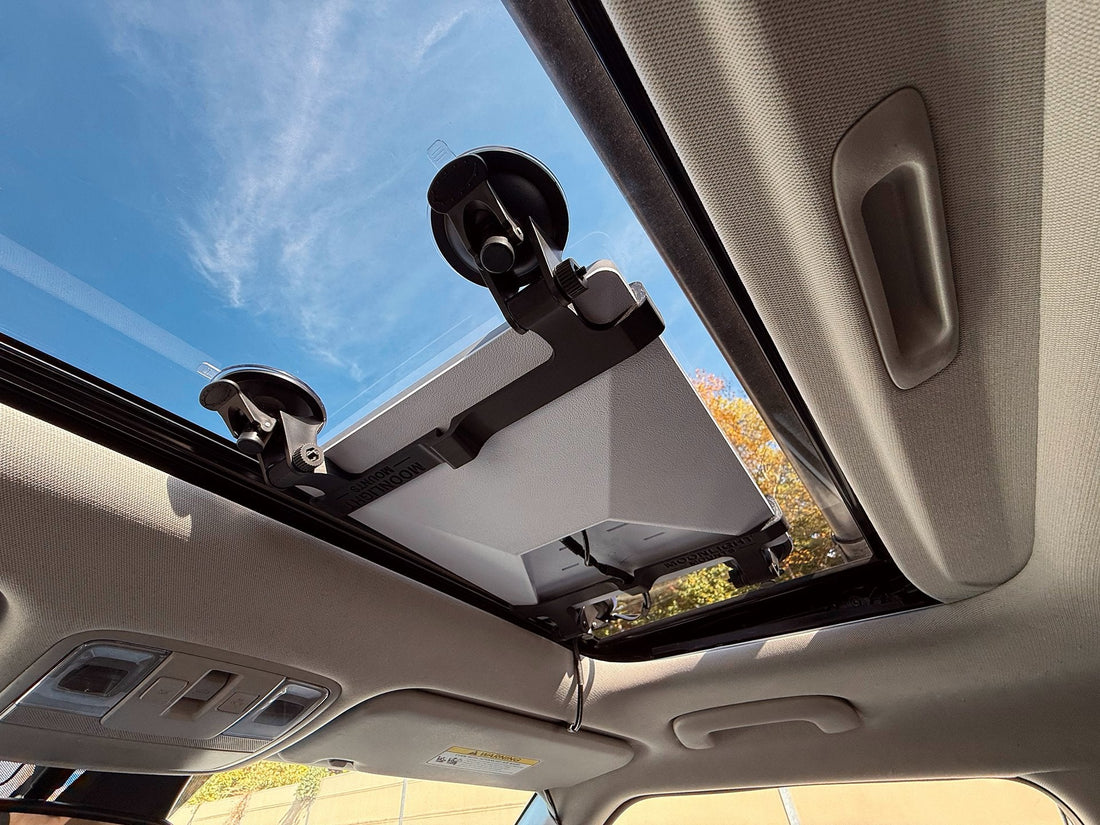 Starlink Mini held against the inside of a car's roof glass by a black suction cup mount, seen from the back seat with blue sky beyond